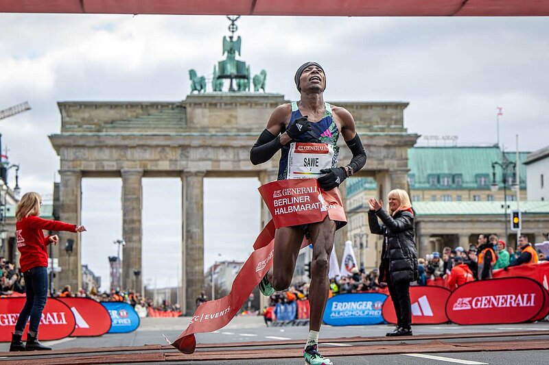 Sebastian Sawe crosses the finish line of the GENERALI BERLIN HALF MARATHON, with the Brandenburg Gate in the background