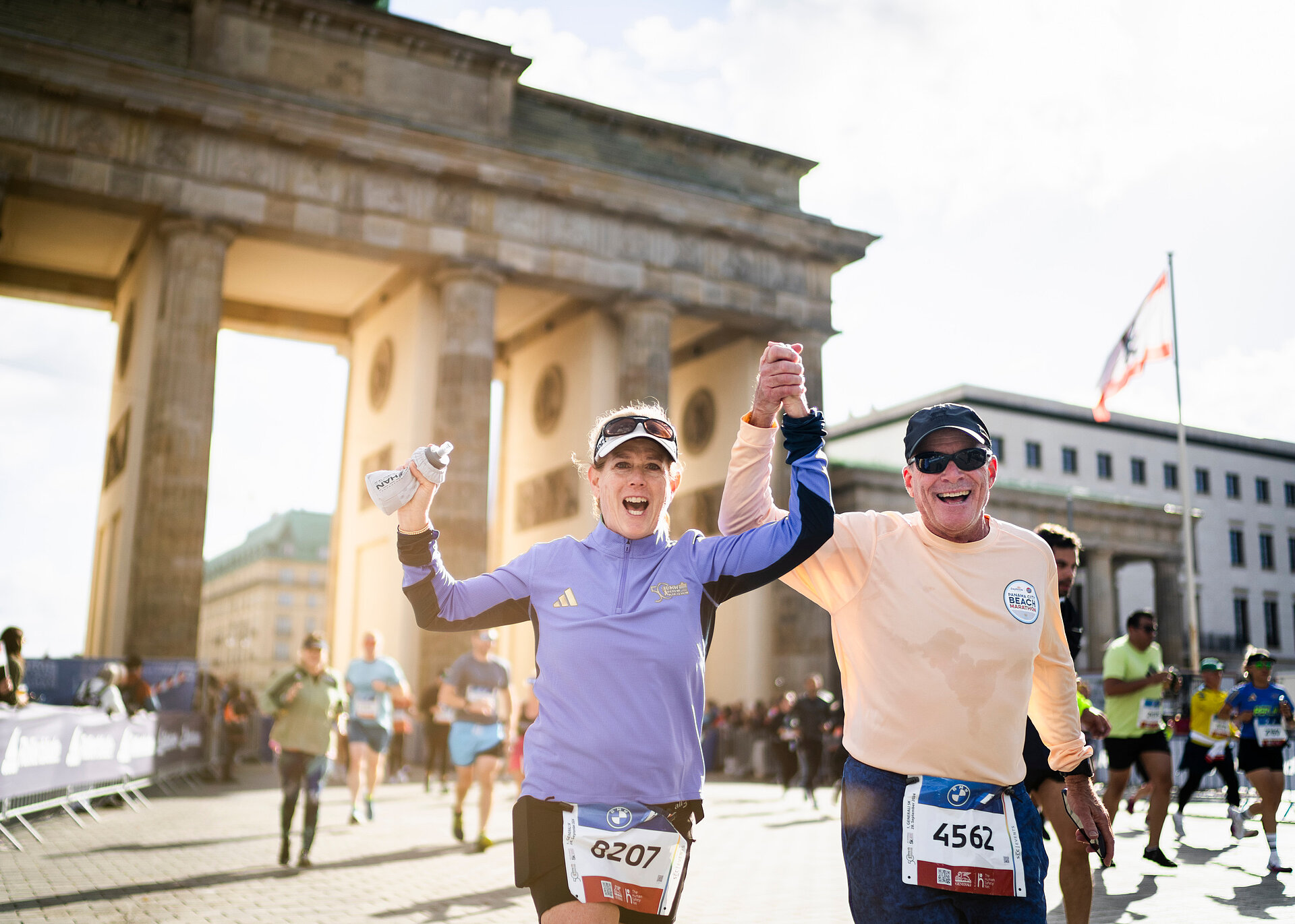 A male runner and a female runner, cheering and holding hands, behind the Brandenburg Gate.